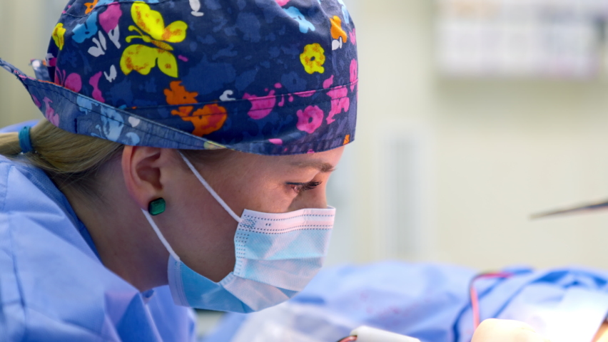 Young blonde female doctor in blur cap and mask working at procedure. Portrait of a surgeon focused on surgical operation. Close up. Royalty-Free Stock Footage #1095550511