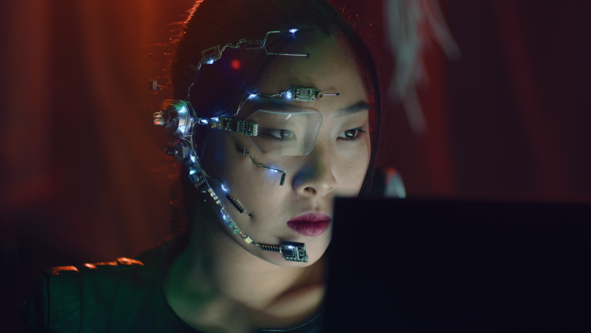 A Cyberpunk girl works on the computer in the red neon lights. Asian girl with futuristic one-eyed glasses and microphone. Cyber and sci-fi backgrounds. Royalty-Free Stock Footage #1095551391