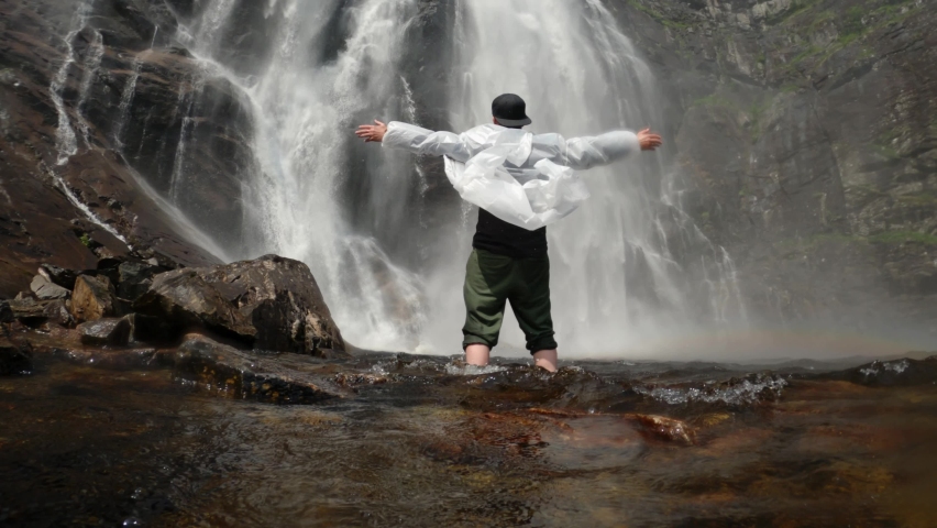 Happy man in a transparent raincoat stands with hands up at the foot of a waterfall in norway near a rock