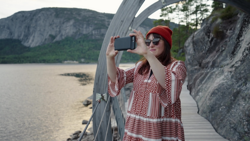 Young girl in a hat is standing on a wooden bridge and taking pictures of the sea on the phone.