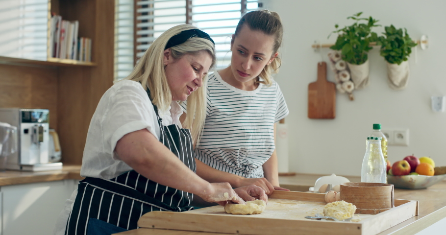 Hapy deligted young daughter helping mother pouring oil on homemade dough while middle aged mother kneading. Women in modern kitchen cooking together having fun while preparing dinner for famly.