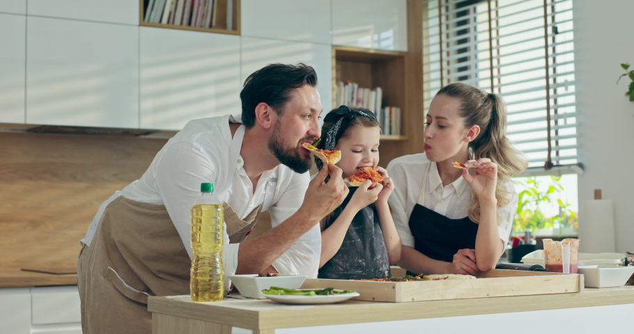Happy family in kitchen cooking baking together spending time at weekeend. Preschooler kid tasting homemade domestic pizza with handsome young father and pretty mother in aprons.
