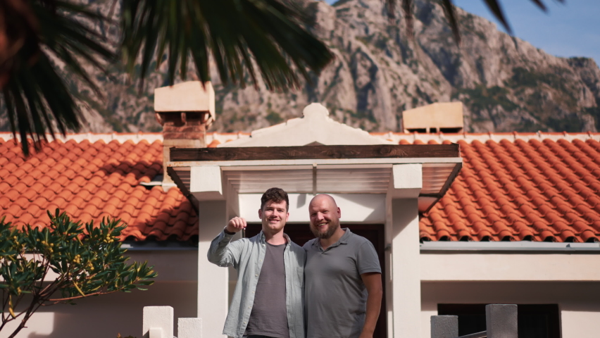 Beautiful young gay couple staying holding a key at background of new house. Two hapy homosexual men showing keys to new apartment lovely area on sunny day. Concept of Pride, lgbt family