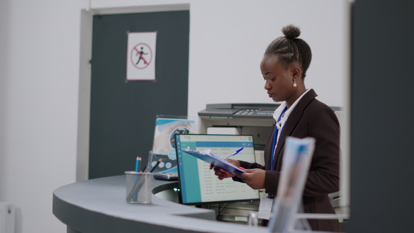 Female receptionist taking notes on papers and using medical report to prepare consultation appointments and checkup visits at reception counter. Hospital worker at registration desk in clinic lobby.
