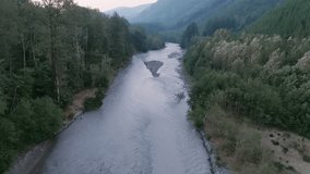 Aerial footage flying over a grey river in between two banks of evergreen trees in the Cascade Mountains in Washington.