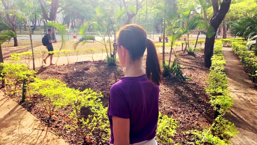 Rear view of young female with ponytail leisurely walking through a park in a city. Real people lifestyle concept. 