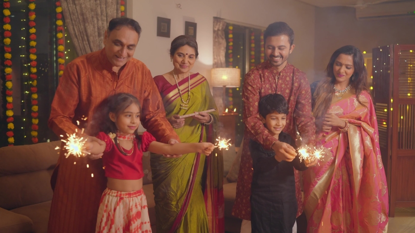 Ethnic Indian Hindu happy smiling cute young boy and a girl or kids in traditional attire playing with firecrackers or sparklers with the help of family elders or parents during Diwali festival. | Shutterstock HD Video #1095567023