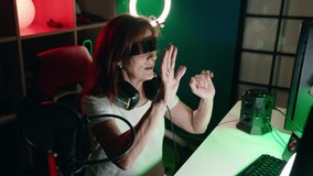 Middle age woman streamer playing video game using virtual reality glasses at gaming room