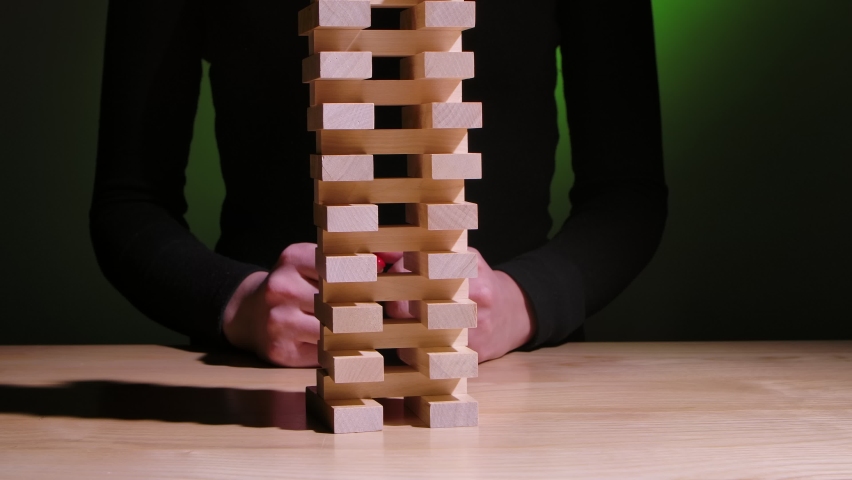 The girl plays the Jenga board game on a green gradient background. The tower of wooden blocks falls when the girl pulls out one of them. Camera movement from right to left. Slow motion. Close up. Royalty-Free Stock Footage #1095574425
