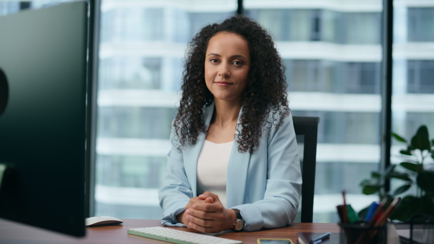Confident young businesswoman looking camera sitting at desk with computer close up. Portrait of attractive latin woman working in modern office wearing blue jacket. Curly lady posing with calm smile. Royalty-Free Stock Footage #1095574665