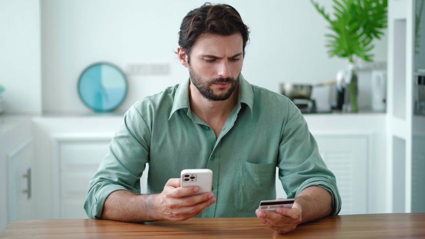 Dark-haired bearded Hispanic young man in casual shirt, using smartphone, transfers money. He is anxious, frustrated not paying the bill due to lack of coins, throws the credit card on a kitchen table Royalty-Free Stock Footage #1095574985