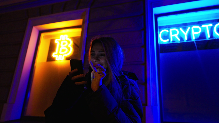 Smiling female crypto investor uses her smartphone buy or sell cryptocurrency at night against bright neon signs BITCOIN, CRYPTO and EURO on windows