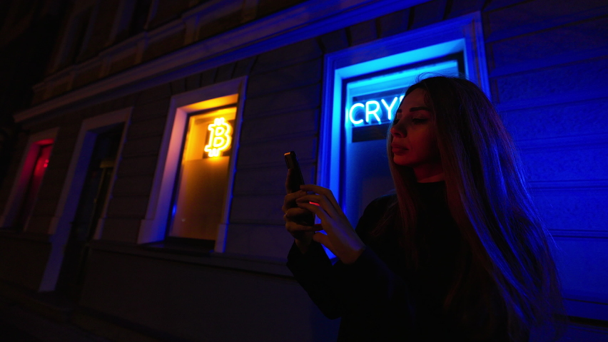 SAD woman after a huge cryptocurrency fall holds her phone against neon Bitcoin and Crypto signs