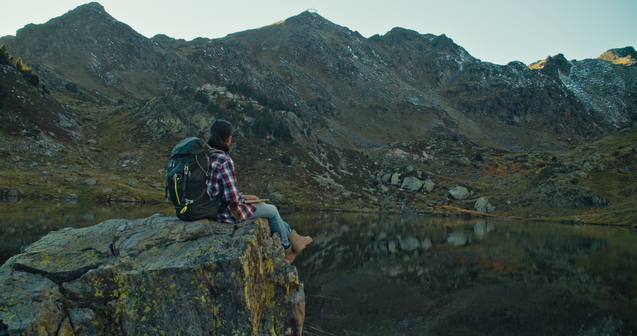 Woman sit on rock at mountain lake with hiker backpack opens notebook computer and make video call wit family or friends. People using technology on vacation travel. 