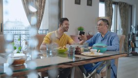 A happy Indian Ethnic young teenage son sitting at the dining table showing a funny video on a mobile phone or a smartphone to his old dad while having lunch or a meal together in an indoor house.