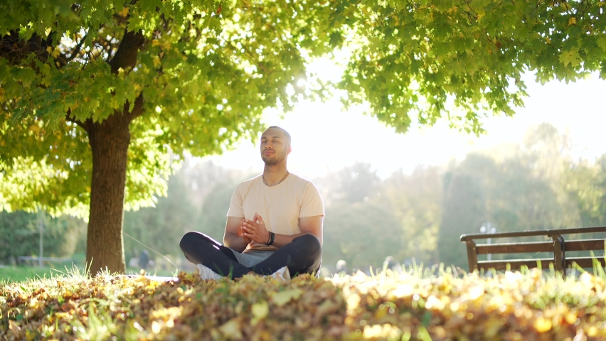 Young athletic man practices yoga in a park or forest in nature, sitting in lotus position on mat in nature. Male meditating relaxing breathing deeply with closed eyes outdoors. Outside. sunlight Royalty-Free Stock Footage #1095697257