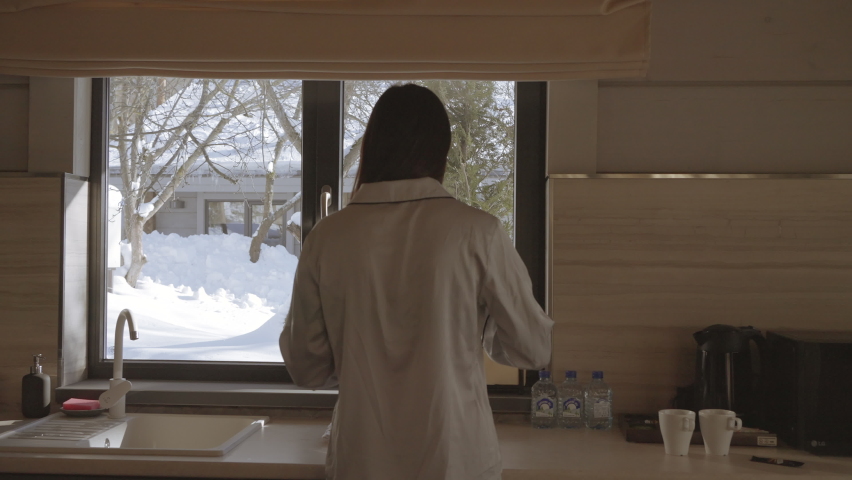 Rear view of a woman making hot beverage in front of the window and winter outside. Action. Cozy kitchen and a girl making morning tea.