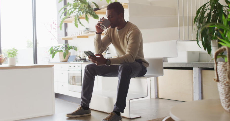 Happy african american man sitting on stairs in kitchen, drinking coffee and using smartphone. Spending quality time at home alone.