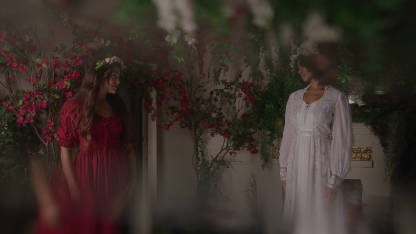 Two Young Women in Dresses Reach Out to Touch Hands in a Dark Garden Room