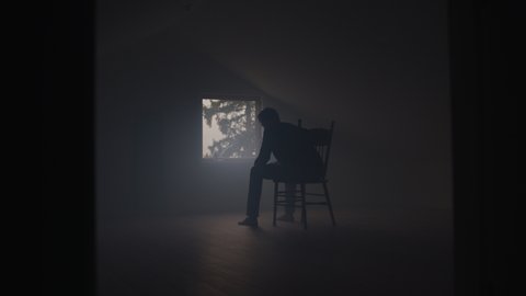 Silhouetted Depressed Young Man Sits on Chair in Smokey Attic in Window Light Adlı Stok Video