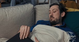 Sick man with beard is lying on sofa and covered with blanket. Man with dark hair is holding laptop and having video chat with family. He tells about well-being. Brunette wipes nose with napkin.