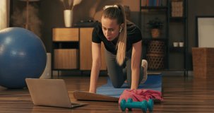 A strong and beautiful fitness girl in sportswear does exercises in her spacious living room with a minimalist interior. She watches online classes on laptop.