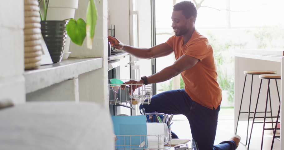 Happy african american man standing in kitchen and loading dishwasher. Spending quality time at home alone. | Shutterstock HD Video #1095748399