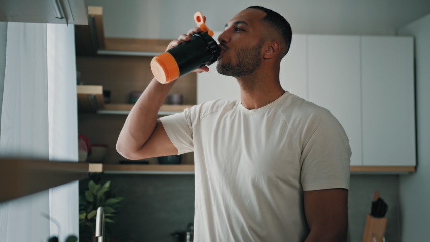 African strong healthy sports man male sport athlete bodybuilder trainer guy in kitchen smile drinking liquid calcium vitamins dietary supplements clean water protein aqua with plastic fitness bottle | Shutterstock HD Video #1095749499