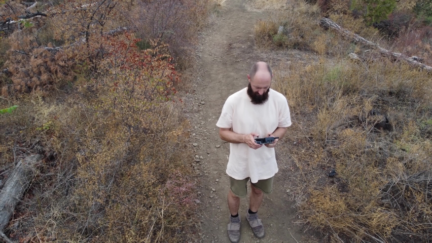 One man with a beard and short hair is flying a drone in the forest on a mountain on a cloudy day. The guy is wearing a beige t-shirt and shorts.