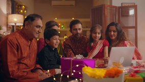 Happy smiling Hindu ethnic Indian close-knit family in traditional attire watching a funny online movie or a comedy video together on a laptop app during Diwali festival season in well decorated home