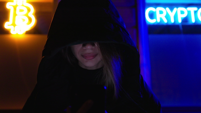 Smiling woman in a hood uses her smartphone to find out the cryptocurrency rise against bright neon signs