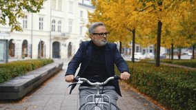 City bike concept. 60-year-old caucasian bearded man dressed in business casual way cycling on rented bike through his favorite park. Slow motion clip. Outdoor shot. High quality 4k footage