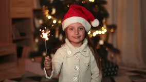 Close up video portrait of lovely cute little girl in warm knitted white sweaters and santa hat holding sparklers in front of decorated Christmas tree, looking at camera.