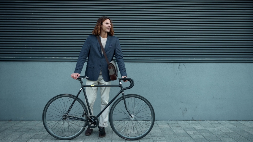 Positive curly haired man wearing jacket looking around while standing with a bicycle outdoors
