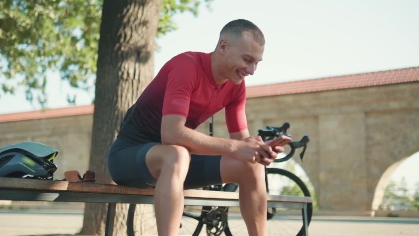 Cheerful sportsman typing on the mobile on the bench near bicycle outdoors