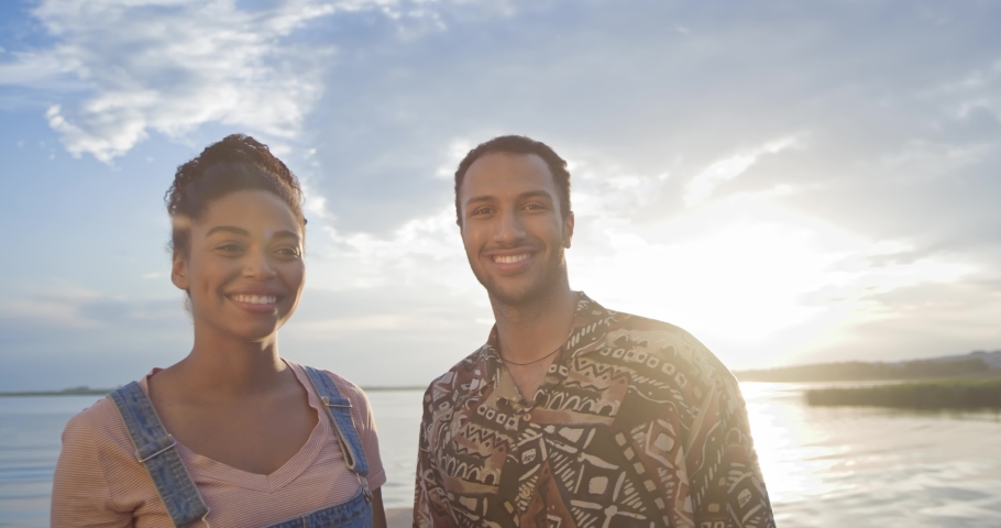 Close up of happy couple man and woman stand on dock near lake looking back and waving hands to friend on boat. African American male and female smiling in good mood. Love relations. Lake concept