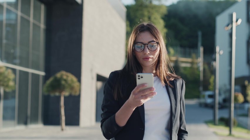 Businesswoman young attractive with glasses walking using the phone in urban city at sunlight look around feel happy. Portrait girl with mobile. Slow motion