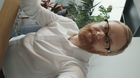 Vertical pov shot of bearded Caucasian businessman in formal shirt and eyeglasses waving and speaking on camera via online video call