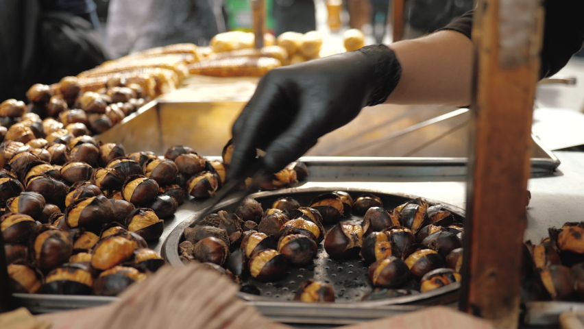 Close up: Delicious Chestnut grilling over glowing coals, make street food in Istanbul, Taksim square. Barbecued roasted on hot stove fresh tasty chestnut, male hand flips  Royalty-Free Stock Footage #1095791715
