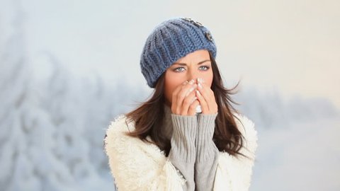 Pretty girl has flu and fever in winter day outdoor. Portrait of beautiful young woman with wool blue cap  looking at camera with snowy landscape behind. 