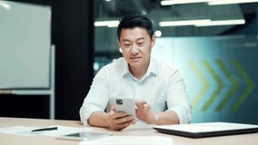 Happy asian office worker sitting at a computer desk in the office using a mobile phone. Cheerful business man entrepreneur employee. typing browses use smartphone reads messages chats communicates - Powered by Shutterstock - Get 15% off with code: PIKWIZARD15