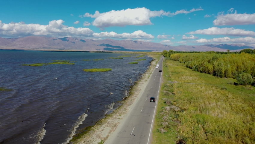 Aerial view highway traffic near water.  Road near Sevan lake in Armenia, Caucasus. Cars driving on asphalt road near lake in Armenia, western Asia. Aerial footage road between lake and field.