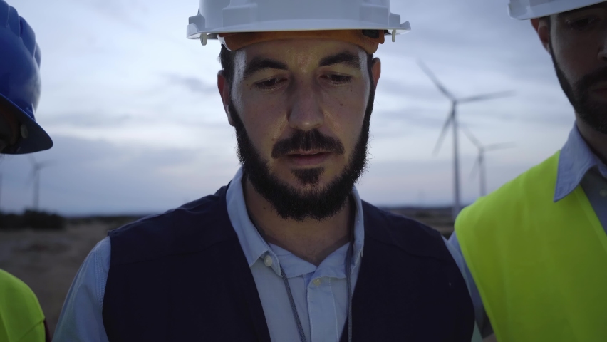 Cinematographic shot of a group of helmeted engineers using an electronic tablet against a background of windmills at sunset. Concept: renewable energy, technology, electricity, utility, green, future Royalty-Free Stock Footage #1095816767