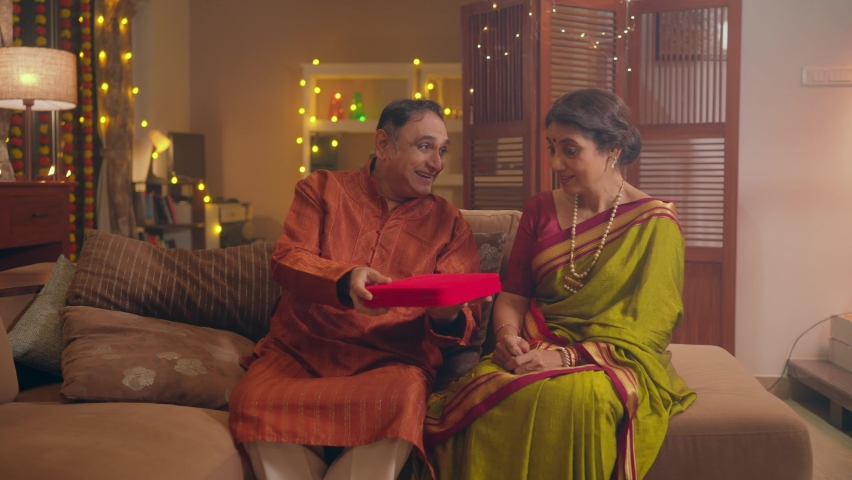 A happy Indian traditional Hindu elderly married couple where an old man or husband surprises his smiling ethnic wife in a saree with a jewellery gift box on the occasion of Diwali festival in a home. Royalty-Free Stock Footage #1095816787
