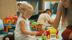 Little white blond girl in dress high-fiving her nursery teacher and playing with toy car with other kids in the background. Horizontal indoor video. High quality 4k footage