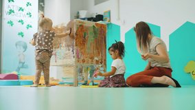 Group of small kids playing around with paint on plastic wrap with their teacher. Creative and messy playtime. Horizontal indoor video. High quality 4k footage