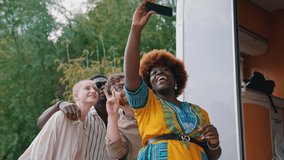 Beautiful smiling black woman in colourful dress with red afro hairstyle taking a selfie with black smartphone with her multiracial friends. Outdoor shot. High quality 4k footage