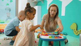 Two young kids boy and girl playing with colorful educational toy with their teacher. Nursery playtime. Multiracial. Horizontal indoor video. High quality 4k footage