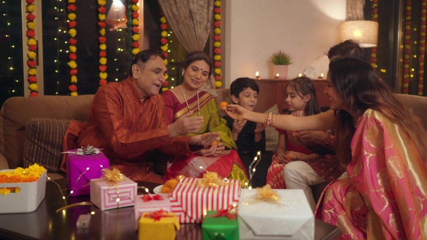 Happy smiling Hindu ethnic Indian family members in traditional Attire eating sweets (Laddoos, Laddus) enjoying the festivities together on the occasion of Diwali festival in a well decorated home. Royalty-Free Stock Footage #1095822899