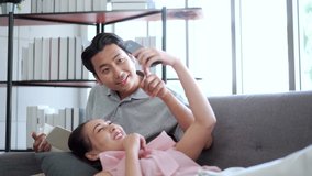 Happy cheerful Asian couple living together in living room in a free time in weekend, Asian girl laying down on sofa with her boyfriend and watching a video on smartphone.